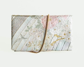 May include: A white clutch bag with a gold chain strap. The bag is decorated with a floral pattern in shades of pink, white, and green.
