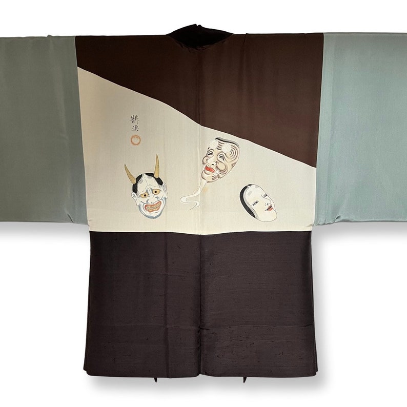 K&ouml;nnte beinhalten: Eine traditionelle japanische Haori-Jacke in Braun und Blau. Die Jacke hat ein wei&szlig;es Feld mit drei bemalten Masken, darunter eine D&auml;monenmaske mit H&ouml;rnern. Der Text "粋" ist in Schwarz auf dem wei&szlig;en Feld geschrieben.