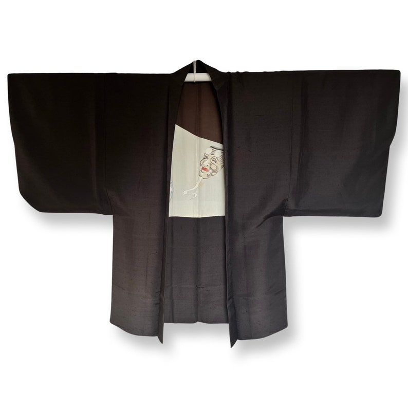 K&ouml;nnte beinhalten: Ein dunkelbraunes japanisches Hanten, eine traditionelle kurze Jacke, mit einem wei&szlig;en Panel, das ein gemaltes Design eines Gesichts zeigt.