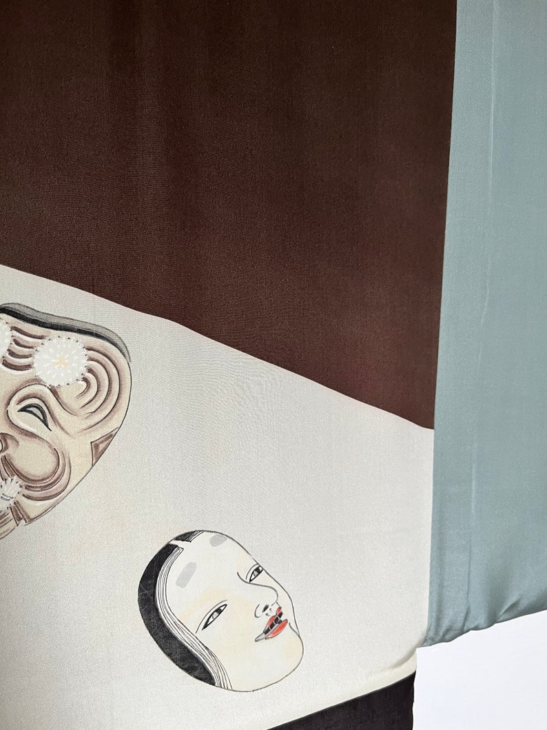 K&ouml;nnte beinhalten: Ein wei&szlig;er Stoff mit zwei schwarz-wei&szlig;en Masken. Die Masken sind auf den Stoff gemalt. Der Stoff ist Teil eines gr&ouml;&szlig;eren Stoffst&uuml;cks mit braunen und blauen Abschnitten.
