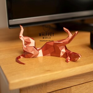 K&ouml;nnte beinhalten: Eine geometrische, kupferfarbene Skulptur eines Hundes in entspannter Pose. Das Kunstwerk wird auf einer Holzoberfl&auml;che pr&auml;sentiert, mit dem Text "WHIPPET WORLD" und einer Website-Adresse. Das Design der Skulptur weist scharfe Winkel und Facetten auf.