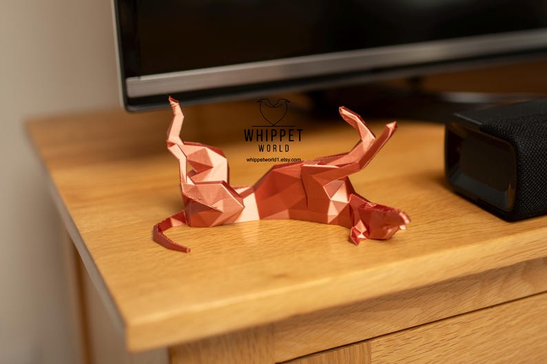 K&ouml;nnte beinhalten: Eine geometrische, kupferfarbene Skulptur eines Hundes in entspannter Pose. Das Kunstwerk wird auf einer Holzoberfl&auml;che pr&auml;sentiert, mit dem Text "WHIPPET WORLD" und einer Website-Adresse. Das Design der Skulptur weist scharfe Winkel und Facetten auf.