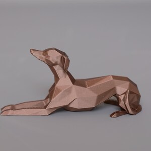 K&ouml;nnte beinhalten: Bronze farbene, geometrische Hundeskulptur in liegender Pose. Das Kunstwerk weist ein Low-Poly-Design auf, wobei abgewinkelte Ebenen die Form eines Hundes bilden. Die Skulptur steht vor einem grauen Hintergrund.