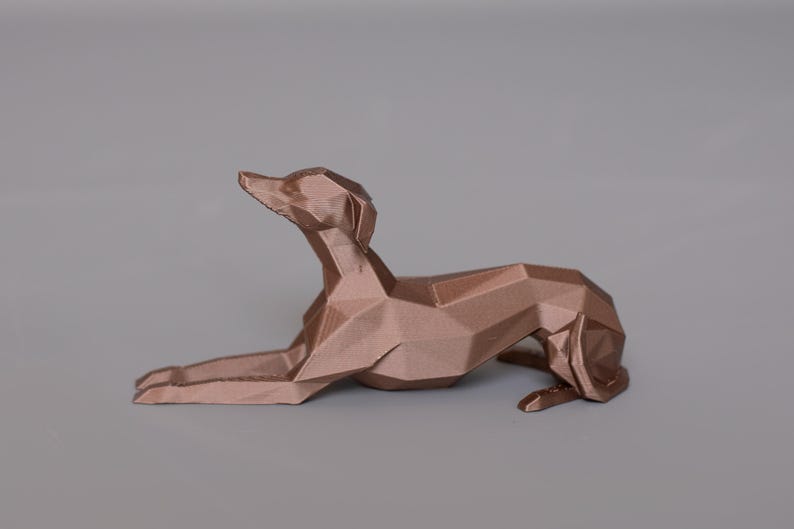 K&ouml;nnte beinhalten: Bronze farbene, geometrische Hundeskulptur in liegender Pose. Das Kunstwerk weist ein Low-Poly-Design auf, wobei abgewinkelte Ebenen die Form eines Hundes bilden. Die Skulptur steht vor einem grauen Hintergrund.