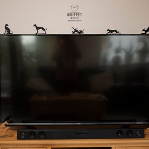 May include: Black metal whippet dog silhouettes in various poses are displayed on top of a large black television. The logo "WHIPPET WORLD" is visible above the TV. A wooden sound bar and decorative motorcycle are on the wooden cabinet below.