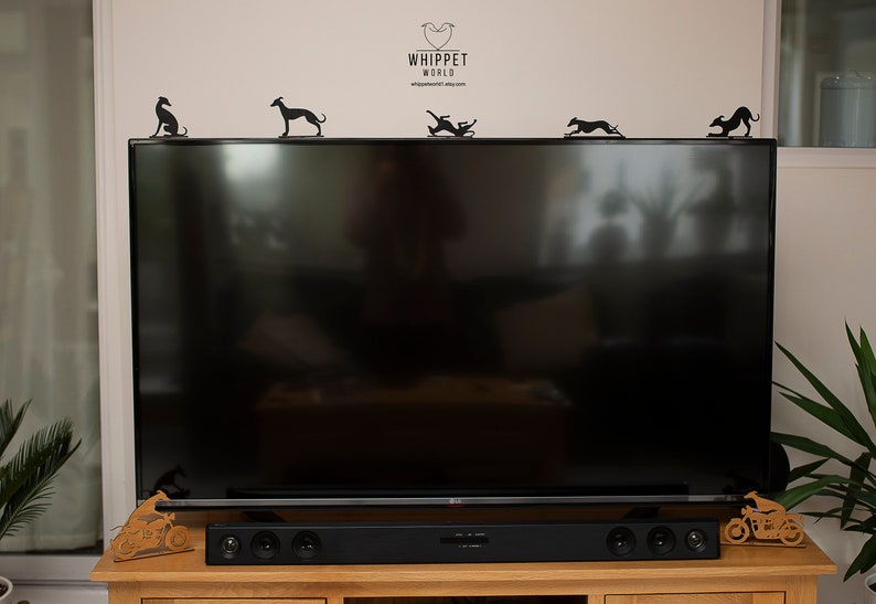 May include: Black metal whippet dog silhouettes in various poses are displayed on top of a large black television. The logo "WHIPPET WORLD" is visible above the TV. A wooden sound bar and decorative motorcycle are on the wooden cabinet below.