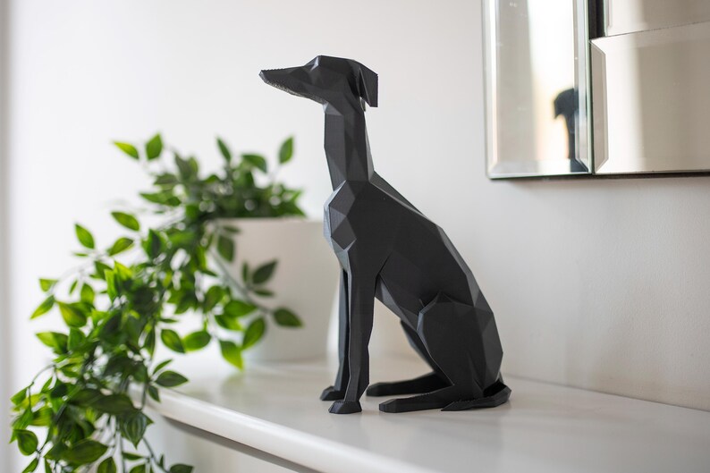 K&ouml;nnte beinhalten: Eine schwarze, geometrische Hundeskulptur mit langem Hals und spitzen Ohren steht auf einem wei&szlig;en Regal. Das moderne Kunstwerk befindet sich neben einem wei&szlig;en Pflanzgef&auml;&szlig; mit gr&uuml;nen Bl&auml;ttern und einem Spiegel.