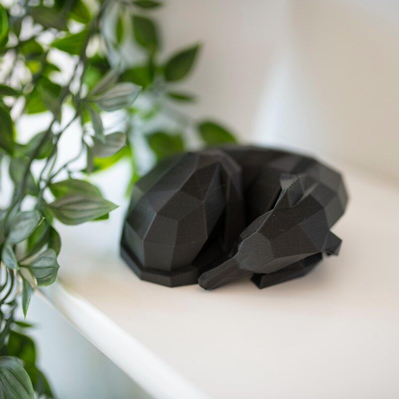 K&ouml;nnte beinhalten: Schwarze geometrische Skulptur eines schlafenden Tieres, m&ouml;glicherweise ein Fuchs oder Hund, ruhend auf einer wei&szlig;en Oberfl&auml;che. Die Skulptur hat ein facettiertes Design und befindet sich in der N&auml;he von gr&uuml;nen Pflanzen. Die Gesamt&auml;sthetik ist modern und minimalistisch.