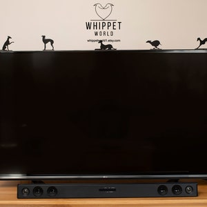 Pode incluir: Uma televis&atilde;o de tela plana preta com uma barra de som preta embaixo. A televis&atilde;o est&aacute; em uma mesa de madeira. H&aacute; v&aacute;rias silhuetas de galgos pretas de metal no topo da televis&atilde;o.