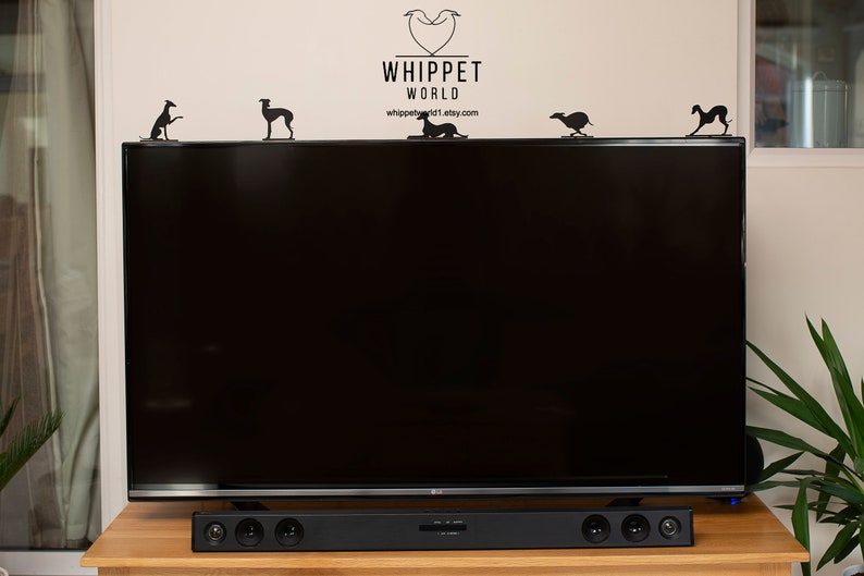 Pode incluir: Uma televis&atilde;o de tela plana preta com uma barra de som preta embaixo. A televis&atilde;o est&aacute; em uma mesa de madeira. H&aacute; v&aacute;rias silhuetas de galgos pretas de metal no topo da televis&atilde;o.