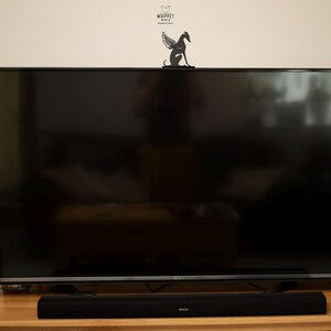 Op de afbeelding: Een grote, zwarte flatscreen-televisie staat op een lichtbruine houten tafel. Een zwarte soundbar is onder het scherm geplaatst. Een decoratieve zwarte metalen whippet met vleugels staat bovenop de tv. De muur boven de tv is wit.