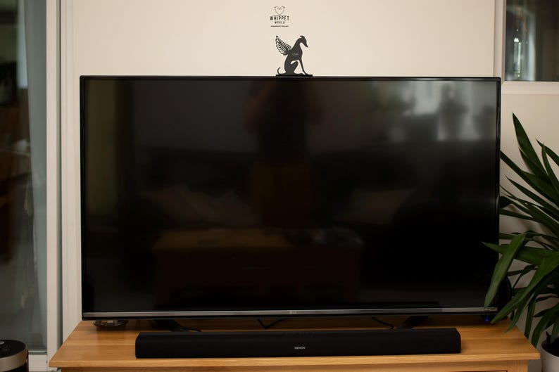 Op de afbeelding: Een grote, zwarte flatscreen-televisie staat op een lichtbruine houten tafel. Een zwarte soundbar is onder het scherm geplaatst. Een decoratieve zwarte metalen whippet met vleugels staat bovenop de tv. De muur boven de tv is wit.