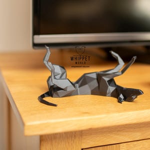 K&ouml;nnte beinhalten: Eine geometrische Papierskulptur eines Windhundes in Graut&ouml;nen, liegend auf einer Holzoberfl&auml;che. Die Skulptur besteht aus gefaltetem Papier und hat ein Low-Poly-Design. Das Logo "WHIPPET WORLD" ist sichtbar.