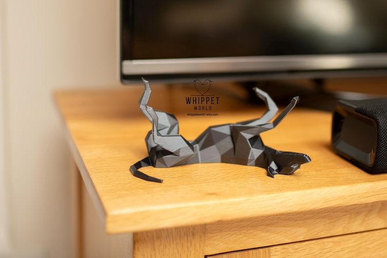 K&ouml;nnte beinhalten: Eine geometrische Papierskulptur eines Windhundes in Graut&ouml;nen, liegend auf einer Holzoberfl&auml;che. Die Skulptur besteht aus gefaltetem Papier und hat ein Low-Poly-Design. Das Logo "WHIPPET WORLD" ist sichtbar.