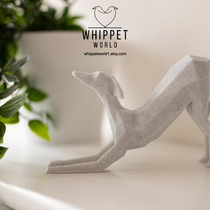 K&ouml;nnte beinhalten: Eine hellgraue, geometrische Whippet-Skulptur in einer Dehnpose. Die Skulptur steht auf einer wei&szlig;en Oberfl&auml;che, mit dem Text "WHIPPET WORLD" und einer Website-Adresse im Hintergrund. Eine gr&uuml;ne Pflanze befindet sich in der linken Ecke.