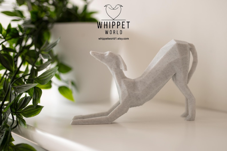 K&ouml;nnte beinhalten: Eine hellgraue, geometrische Whippet-Skulptur in einer Dehnpose. Die Skulptur steht auf einer wei&szlig;en Oberfl&auml;che, mit dem Text "WHIPPET WORLD" und einer Website-Adresse im Hintergrund. Eine gr&uuml;ne Pflanze befindet sich in der linken Ecke.