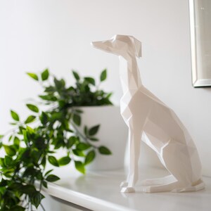 K&ouml;nnte beinhalten: Eine wei&szlig;e, geometrische Hundeskulptur sitzt auf einer wei&szlig;en Oberfl&auml;che, mit einer gr&uuml;nen Pflanze in einem wei&szlig;en Topf links. Der Hund hat einen langen Hals und spitze Ohren. Ein Spiegel befindet sich im Hintergrund.