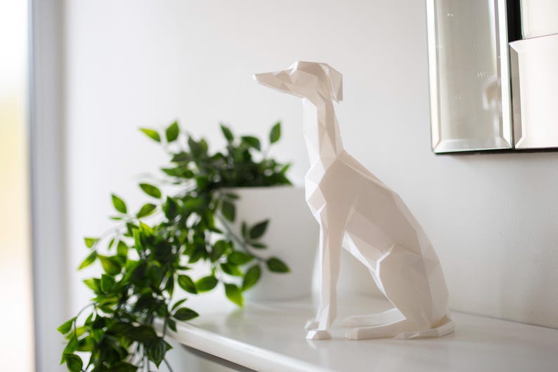K&ouml;nnte beinhalten: Eine wei&szlig;e, geometrische Hundeskulptur sitzt auf einer wei&szlig;en Oberfl&auml;che, mit einer gr&uuml;nen Pflanze in einem wei&szlig;en Topf links. Der Hund hat einen langen Hals und spitze Ohren. Ein Spiegel befindet sich im Hintergrund.