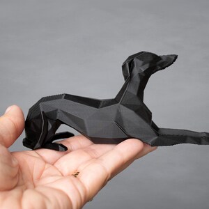 K&ouml;nnte beinhalten: Eine schwarze, geometrische Hundeskulptur in liegender Pose. Der Hund besteht aus eckigen, facettierten Formen, was ihm eine moderne, minimalistische &Auml;sthetik verleiht. Die Skulptur wird in einer Hand vor grauem Hintergrund gehalten.