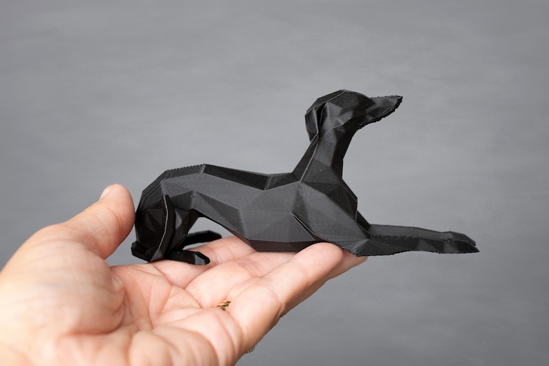 K&ouml;nnte beinhalten: Eine schwarze, geometrische Hundeskulptur in liegender Pose. Der Hund besteht aus eckigen, facettierten Formen, was ihm eine moderne, minimalistische &Auml;sthetik verleiht. Die Skulptur wird in einer Hand vor grauem Hintergrund gehalten.