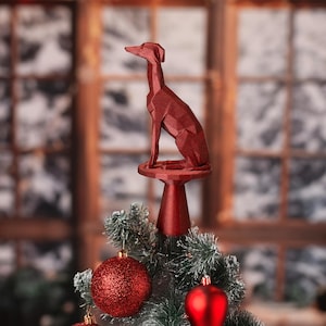 May include: A red geometric dog Christmas tree topper. The dog is perched atop a small pedestal, and the tree is decorated with red ornaments. The background is a blurred view of a window.