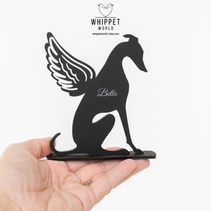 Op de afbeelding: Zwart metalen whippet hond silhouet met engelenvleugels, gepersonaliseerd met de naam "Bella". De hond zit, met vleugels op zijn rug. Het Whippet World-logo staat in de linkerbovenhoek.