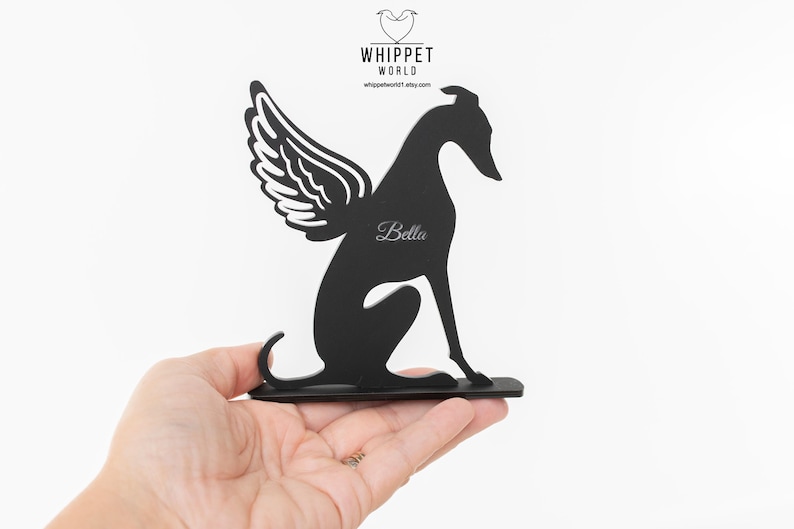 Op de afbeelding: Zwart metalen whippet hond silhouet met engelenvleugels, gepersonaliseerd met de naam "Bella". De hond zit, met vleugels op zijn rug. Het Whippet World-logo staat in de linkerbovenhoek.