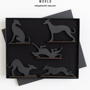 May include: A black box contains five wooden whippet dog silhouettes in various poses. The dogs are dark gray and mounted on small, brown wooden bases. The box is black with a black lid and sits on a white surface. The top of the box has the text "WHIPPET WORLD".