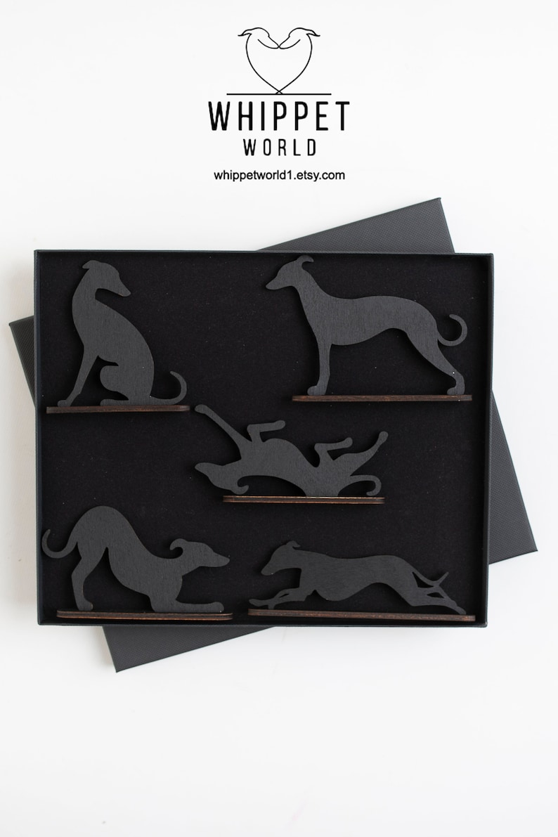 May include: A black box contains five wooden whippet dog silhouettes in various poses. The dogs are dark gray and mounted on small, brown wooden bases. The box is black with a black lid and sits on a white surface. The top of the box has the text "WHIPPET WORLD".
