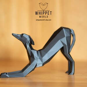 K&ouml;nnte beinhalten: Eine geometrische, graue Skulptur eines Hundes in einer Dehnpose. Die Skulptur besteht aus winkligen Ebenen, die ihr ein modernes, abstraktes Aussehen verleihen. Das Logo "WHIPPET WORLD" und die Website-Adresse sind sichtbar.