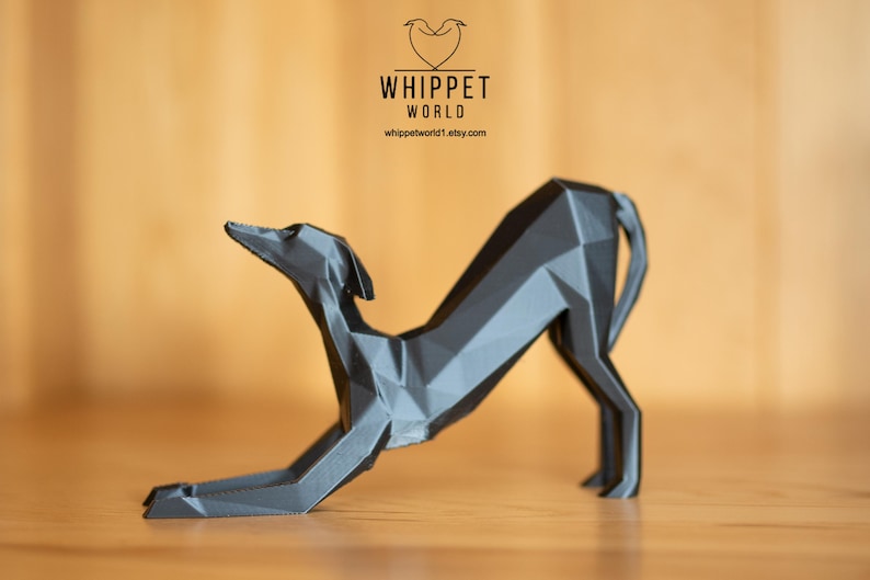 K&ouml;nnte beinhalten: Eine geometrische, graue Skulptur eines Hundes in einer Dehnpose. Die Skulptur besteht aus winkligen Ebenen, die ihr ein modernes, abstraktes Aussehen verleihen. Das Logo "WHIPPET WORLD" und die Website-Adresse sind sichtbar.