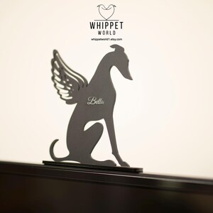 Op de afbeelding: Een donkergrijze metalen whippet-hondsilhouet met engelen vleugels. De naam van de hond, "Bella", is op het lichaam gegraveerd. Het Whippet World-logo staat bovenaan. De hond zit op een zwart oppervlak.