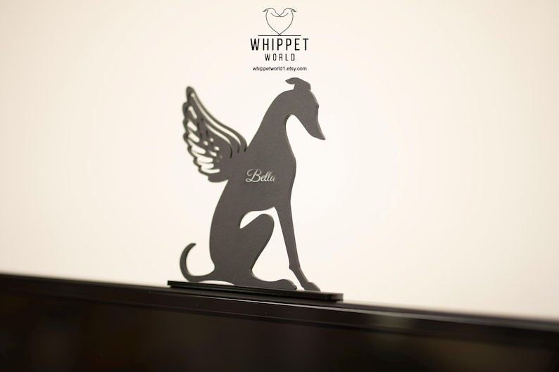 Op de afbeelding: Een donkergrijze metalen whippet-hondsilhouet met engelen vleugels. De naam van de hond, "Bella", is op het lichaam gegraveerd. Het Whippet World-logo staat bovenaan. De hond zit op een zwart oppervlak.