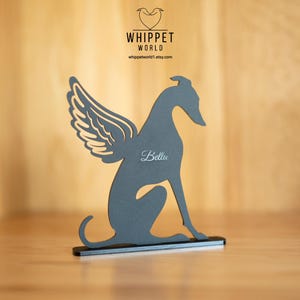 Op de afbeelding: Een grijze silhouet van een whippet-hond met engelen vleugels, zittend op een zwarte basis. De naam van de hond, "Bella", is in cursief geschreven. Het Whippet World-logo en websiteadres zijn zichtbaar op de achtergrond.