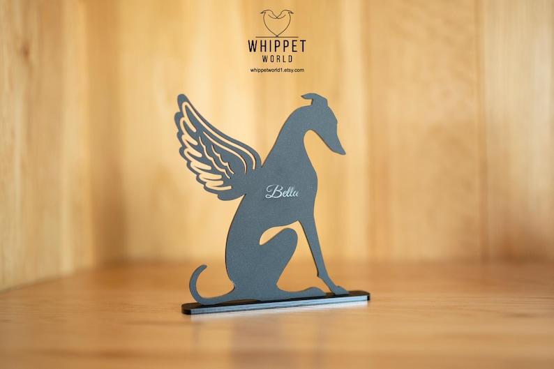 Op de afbeelding: Een grijze silhouet van een whippet-hond met engelen vleugels, zittend op een zwarte basis. De naam van de hond, "Bella", is in cursief geschreven. Het Whippet World-logo en websiteadres zijn zichtbaar op de achtergrond.