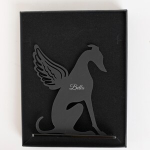 Op de afbeelding: Zwart metalen silhouet van een hond met engelen vleugels, gepersonaliseerd met de naam "Bella". Het kunstwerk wordt gepresenteerd in een zwarte doos, met het "Whippet World" logo en website adres bovenaan.