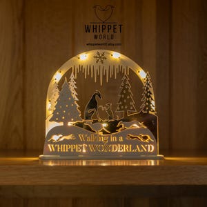 May include: A decorative wooden light-up sign with a winter scene featuring whippets and pine trees. The sign reads "Walking in a Whippet Wonderland" and has a warm, inviting glow. The sign is on a wooden surface.