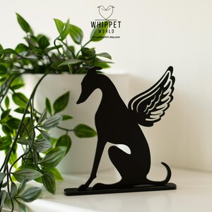 Op de afbeelding: Zwart metalen silhouet van een whippet-hond met engelen vleugels. De hond zit. De afbeelding bevat een witte pot met groene planten en de tekst "WHIPPET WORLD".