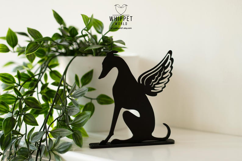 Op de afbeelding: Zwart metalen silhouet van een whippet-hond met engelen vleugels. De hond zit. De afbeelding bevat een witte pot met groene planten en de tekst "WHIPPET WORLD".