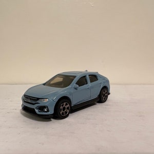 May include: A light blue die-cast model car with black wheels and a black undercarriage. The car has a detailed front grille and headlights. The model is a compact hatchback.