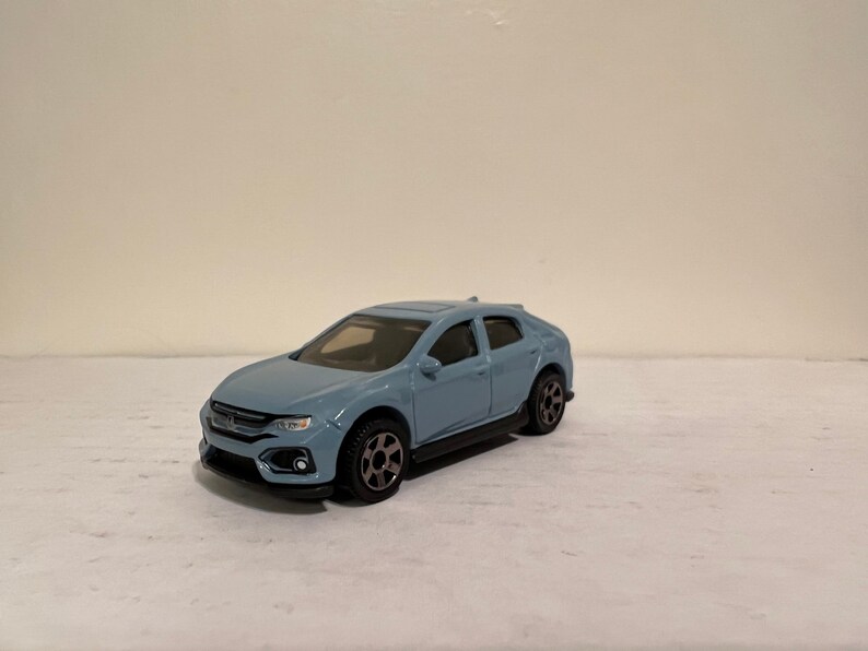 May include: A light blue die-cast model car with black wheels and a black undercarriage. The car has a detailed front grille and headlights. The model is a compact hatchback.