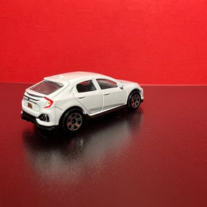 May include: A white die-cast model car with black wheels and a detailed design. The toy car is set against a red background, showcasing its sleek, modern aesthetic. The car has a license plate.
