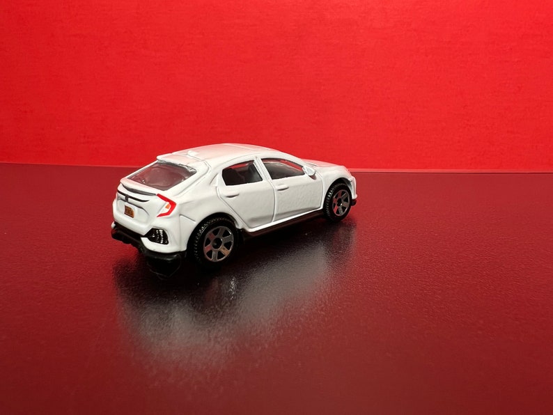 May include: A white die-cast model car with black wheels and a detailed design. The toy car is set against a red background, showcasing its sleek, modern aesthetic. The car has a license plate.