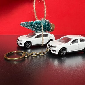 May include: Two white toy cars, one with a small Christmas tree tied to the roof, and the other attached to a keychain. The cars are set against a red background, suggesting a holiday theme. The keychain is made of brass.