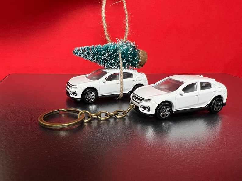 May include: Two white toy cars, one with a small Christmas tree tied to the roof, and the other attached to a keychain. The cars are set against a red background, suggesting a holiday theme. The keychain is made of brass.