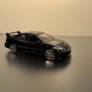 May include: A black die-cast model car with a spoiler and sunroof. The miniature vehicle has silver wheels and is displayed on a reflective surface. The car is a coupe.