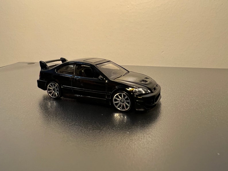 May include: A black die-cast model car with a spoiler and sunroof. The miniature vehicle has silver wheels and is displayed on a reflective surface. The car is a coupe.