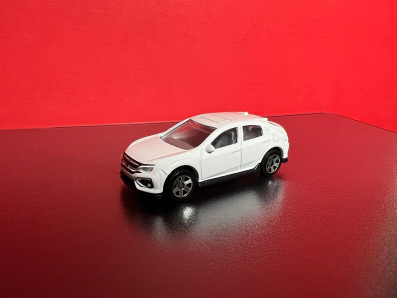 May include: A white toy car with black wheels and a detailed design. The car is set against a red background, creating a striking contrast. The toy car is a miniature replica of a modern vehicle.