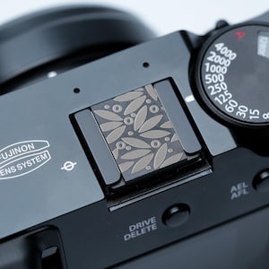 Op de afbeelding: Close-up van een zwarte camera met een zilveren bladerenpatroon op de flitsschoen. De camera heeft een draaiknop met cijfers en instellingen, en de tekst "FUJINON LENS SYSTEM" is zichtbaar. De camera heeft ook de tekst "DRIVE DELETE".