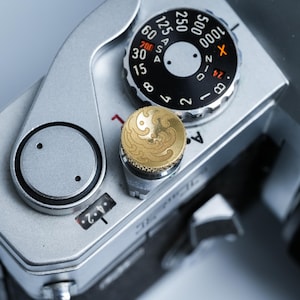 May include: Close-up of a silver film camera with black dials and a gold-colored button. The dials display numbers and settings for photography. The camera's design is classic and detailed, with a focus on its mechanical components.